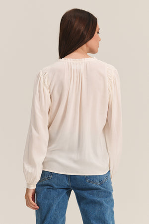 A woman with straight brown hair is shown from the back against a neutral background, wearing the TAMARA BLOUSE by Velvet by Graham & Spencer—a long-sleeve, feminine top with a ruffled neckline—paired with blue jeans.