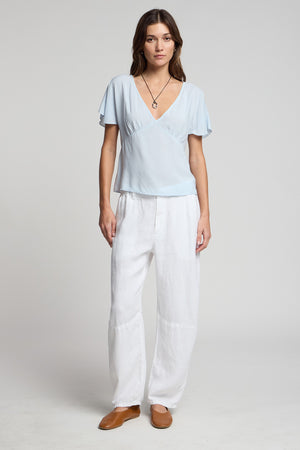 A woman faces forward wearing the light blue LINA TOP by Velvet by Graham & Spencer in soft rayon challis, paired with white loose pants, brown shoes, and a necklace, showcasing a relaxed, feminine silhouette against a plain light background.