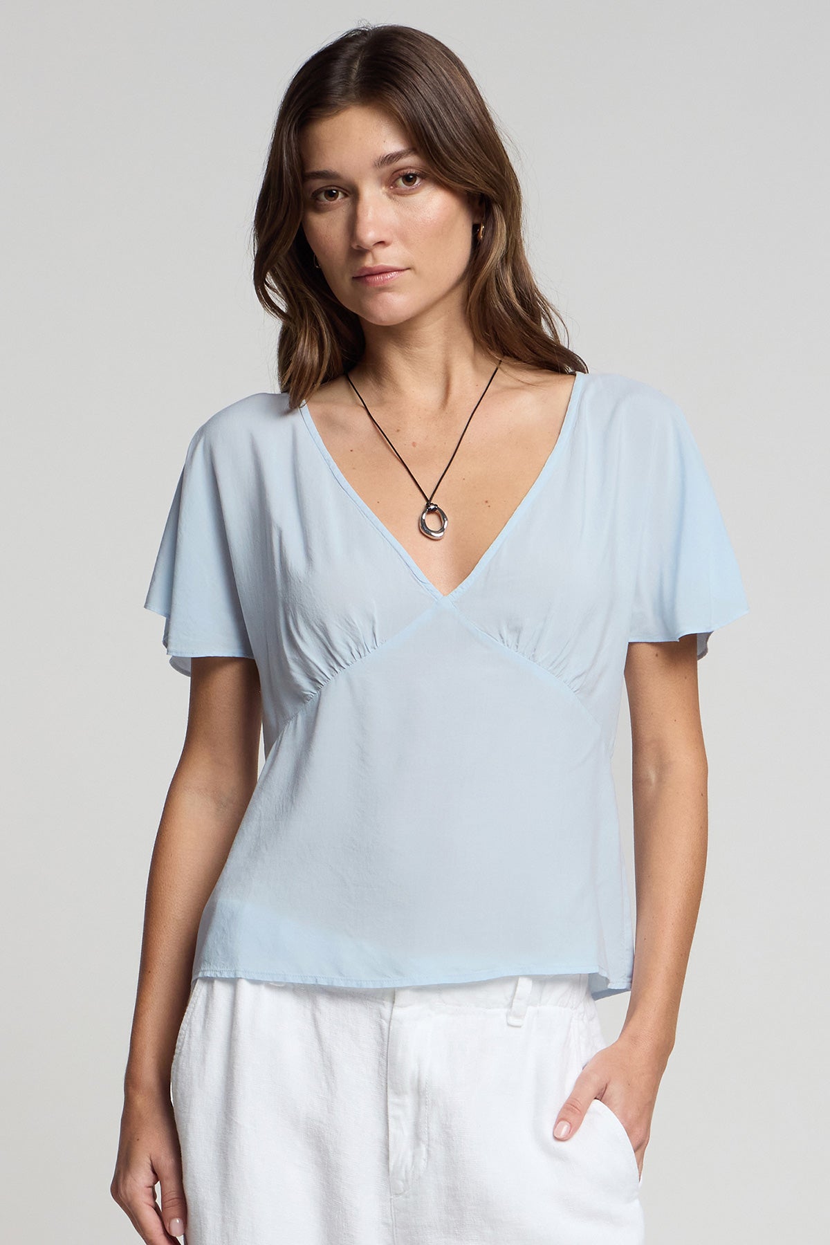 A woman stands against a plain background, wearing the LINA TOP by Velvet by Graham & Spencer in light blue rayon challis, paired with white pants and a simple pendant necklace to highlight her feminine silhouette.-41371958902977