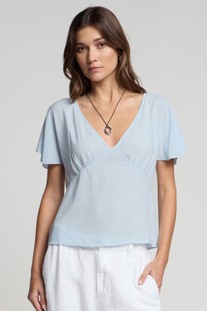A woman stands against a plain background, wearing the LINA TOP by Velvet by Graham & Spencer in light blue rayon challis, paired with white pants and a simple pendant necklace to highlight her feminine silhouette.