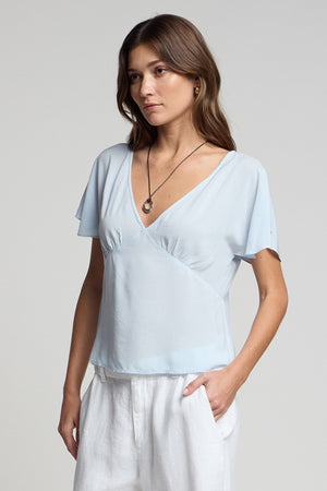 A woman wearing the Velvet by Graham & Spencer LINA TOP in light blue rayon challis and white pants stands with one hand in her pocket, highlighting a feminine silhouette against a plain background.