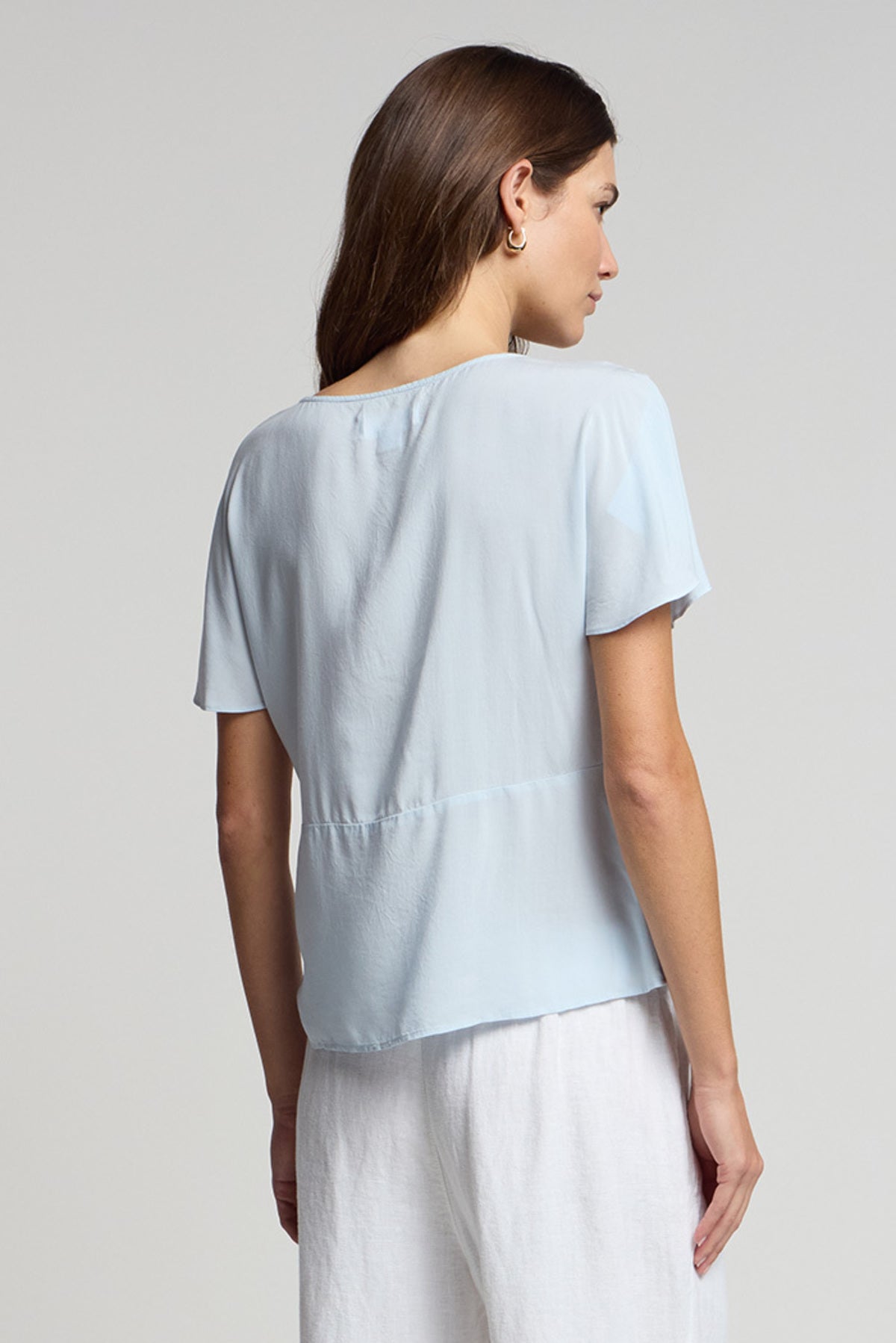 A woman faces away, wearing the light blue LINA TOP by Velvet by Graham & Spencer in rayon challis with white pants, showcasing a soft, feminine silhouette against a simple background.