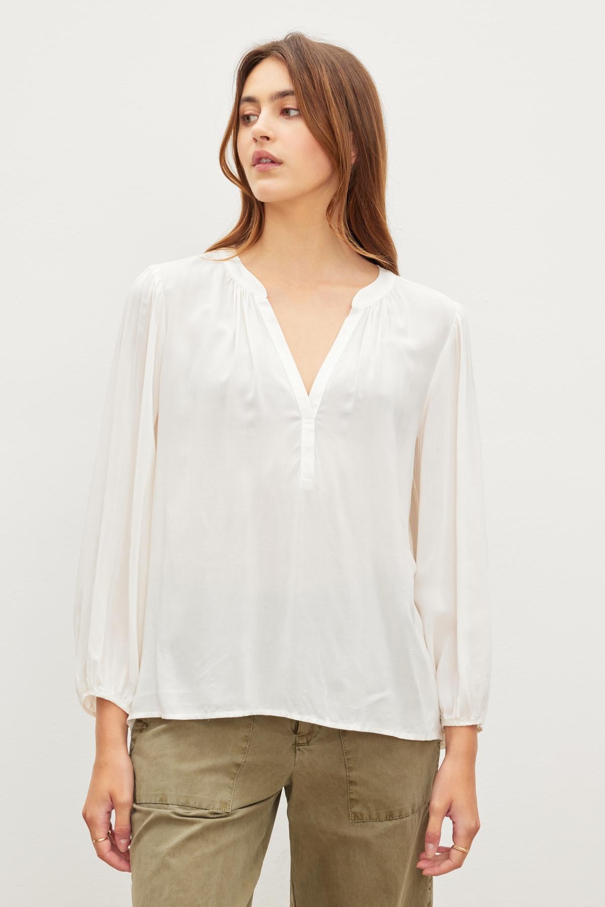 A person with long hair is wearing a white relaxed fit, v-neck ASHLEY TOP by Velvet by Graham & Spencer and olive green pants made from rayon challis while looking to the side.-37229048496321