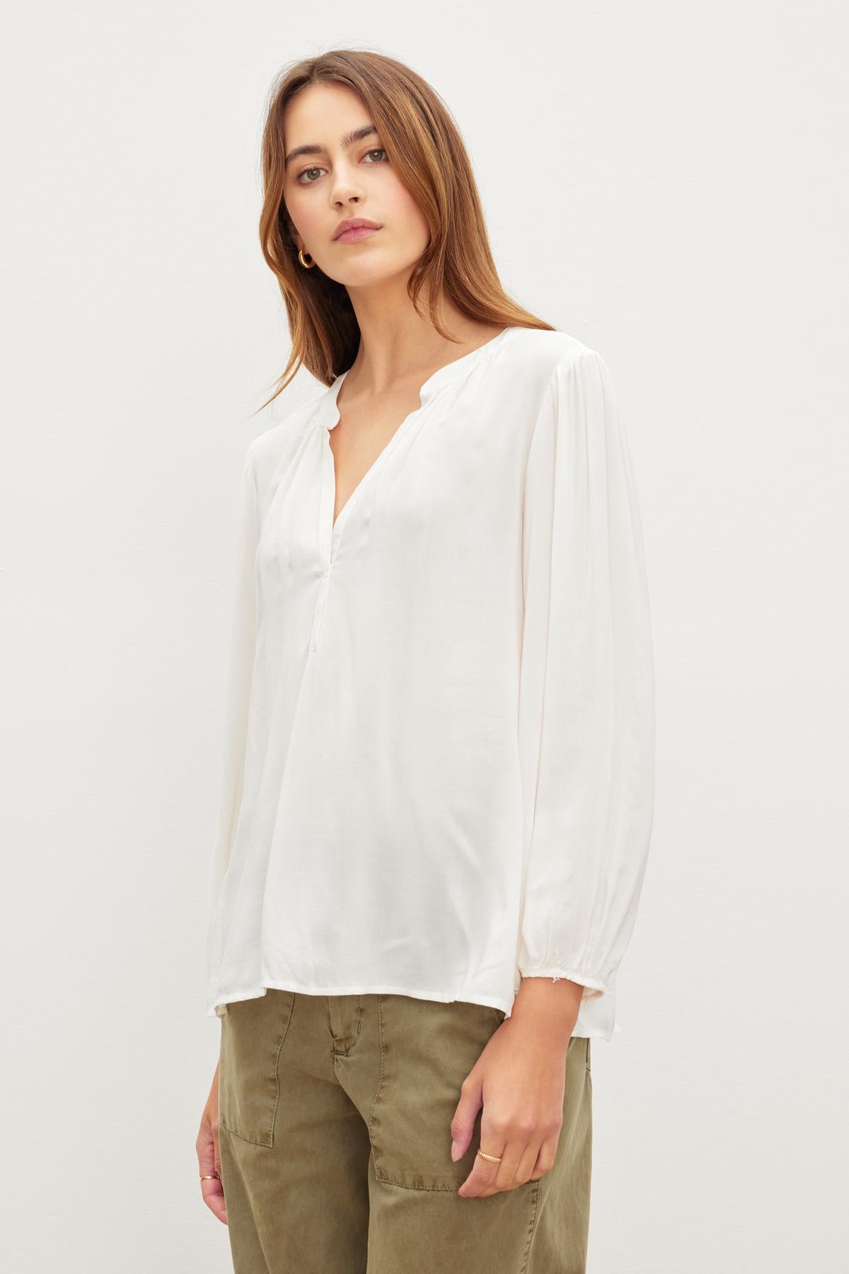   Person wearing a white, long-sleeve ASHLEY TOP by Velvet by Graham & Spencer and green pants stands against a plain white background. 