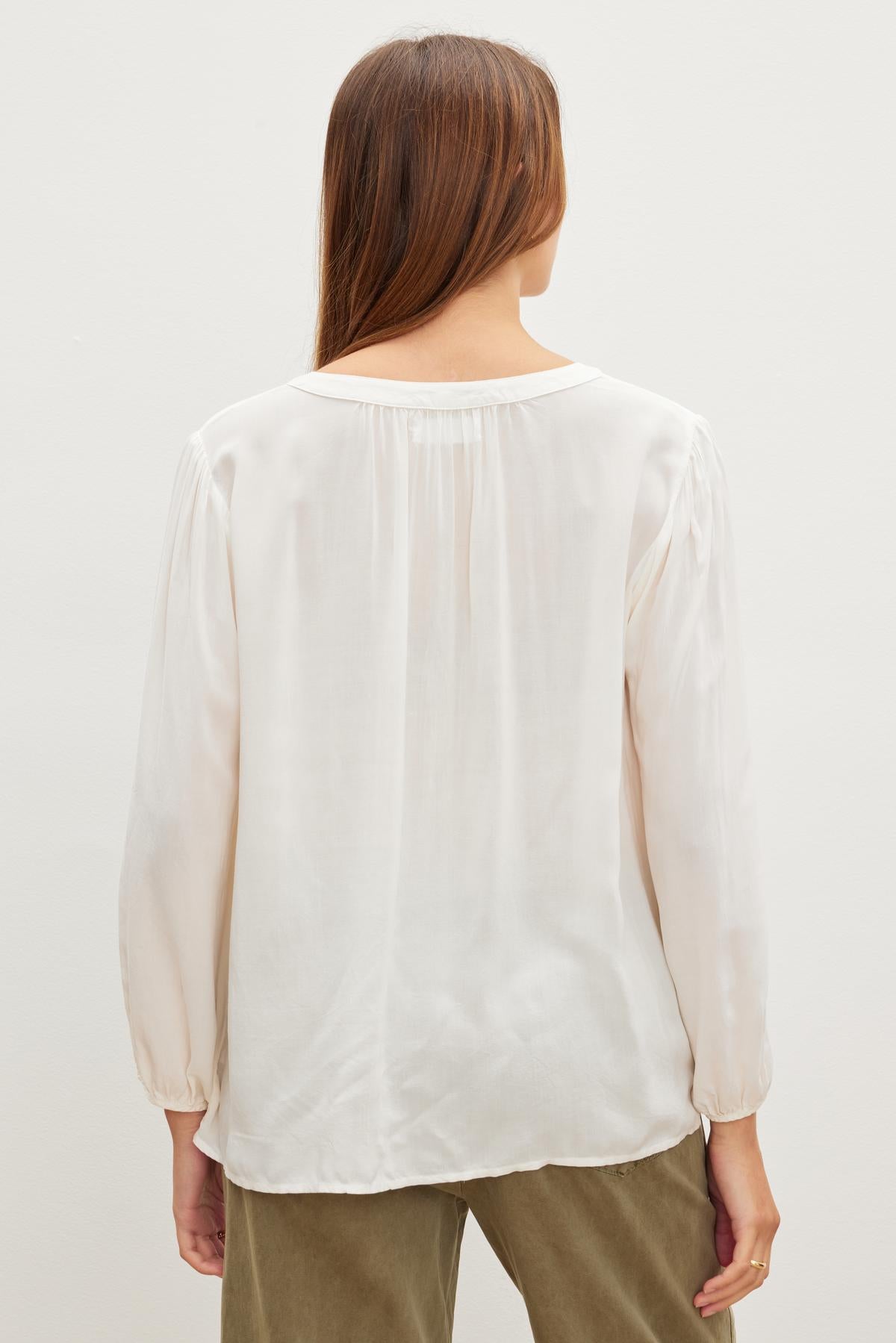   A person with long brown hair is seen from the back, wearing a relaxed fit, ASHLEY TOP by Velvet by Graham & Spencer in a loose-fitting, long-sleeve white blouse style and green pants. 