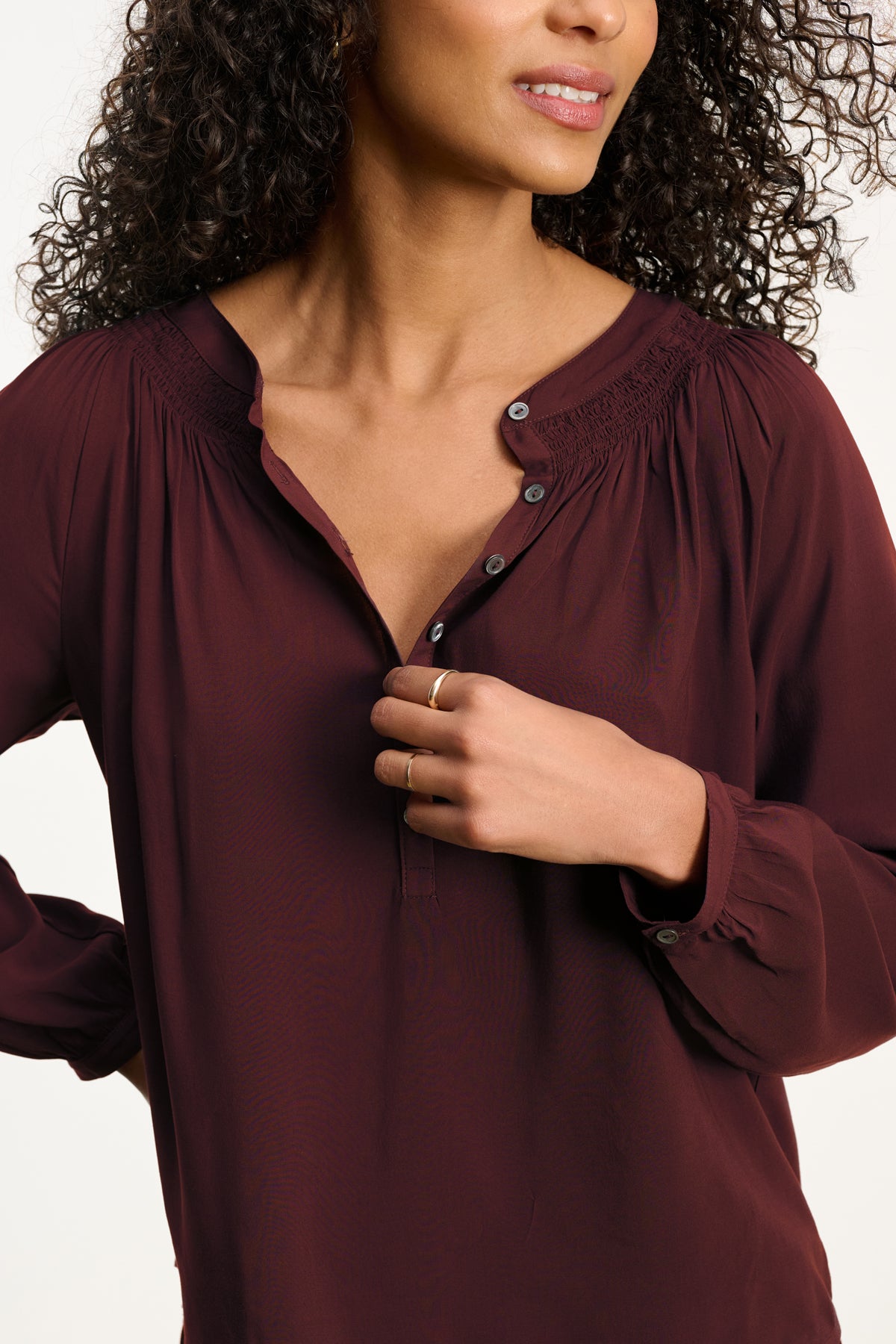 A woman with curly hair, her face partially visible, touches the collar of the maroon AURELIA TOP by Velvet by Graham & Spencer—a relaxed fit, long-sleeve blouse with buttons.-Hover Image