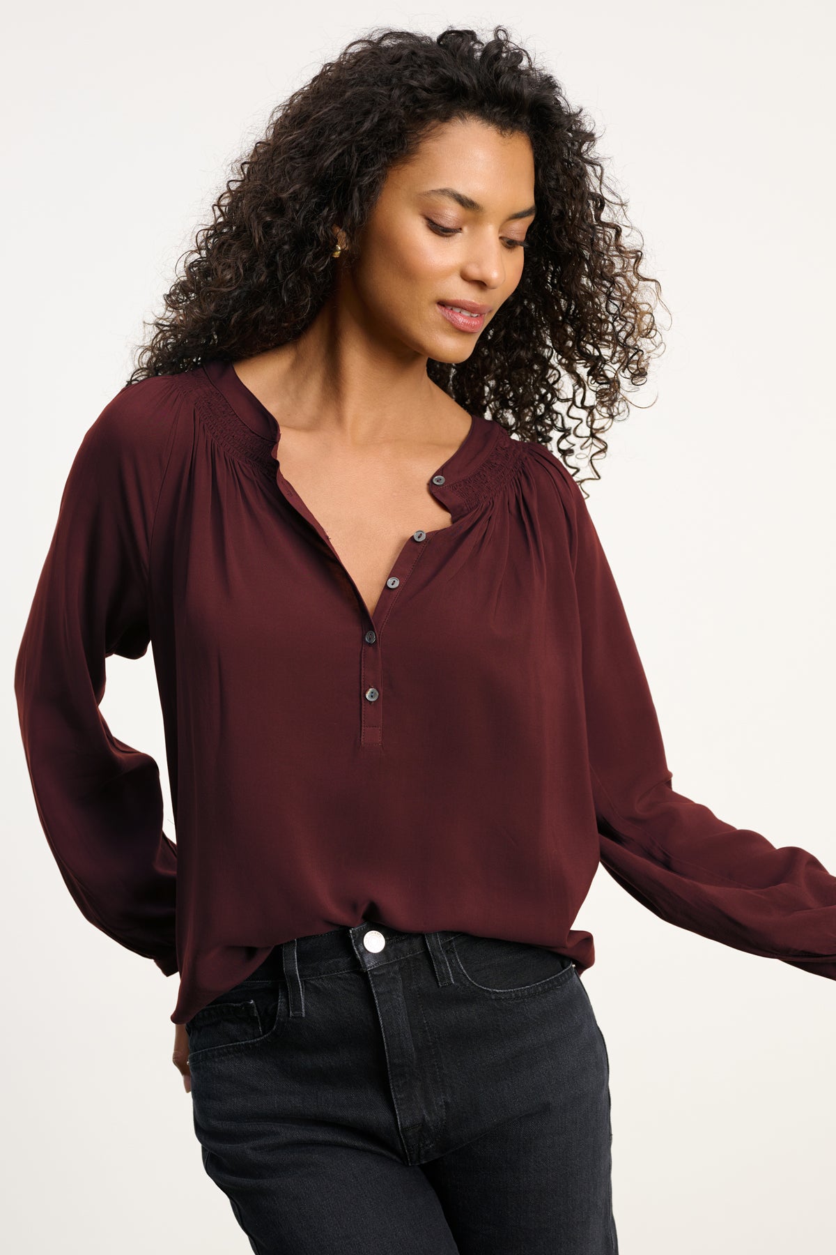 A woman wears the AURELIA TOP by Velvet by Graham & Spencer—a long-sleeve, burgundy blouse with smocked details and button accents—paired with black jeans. The relaxed rayon challis fit ensures comfort against a plain light background.