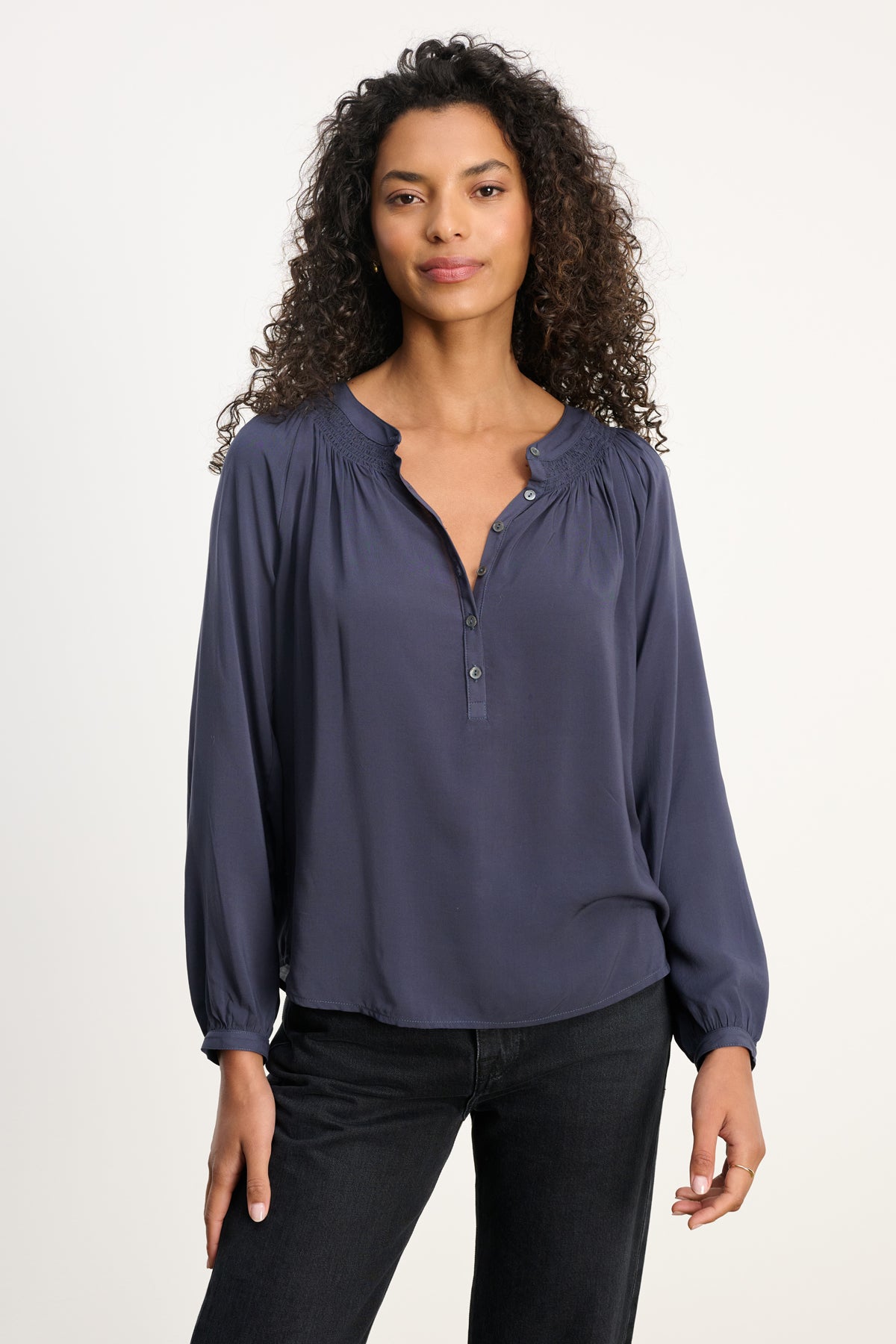   A woman with curly hair wears the Velvet by Graham & Spencer AURELIA TOP, a navy long sleeve made from breathable rayon challis, paired with black pants against a plain light background. 