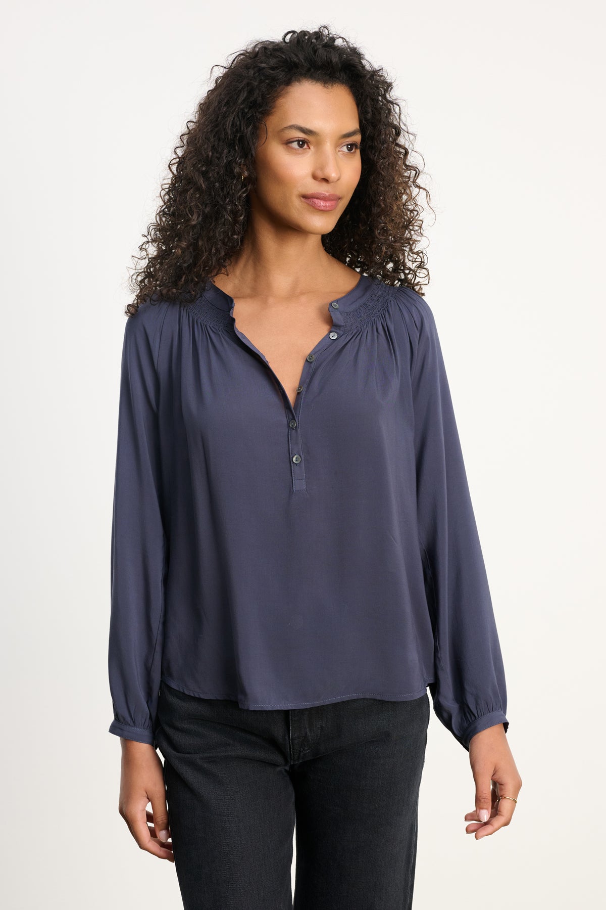 A woman with curly hair wears the Velvet by Graham & Spencer AURELIA TOP, a loose-fit rayon challis blouse with a smocked neckline, paired with black jeans, standing against a plain light background.