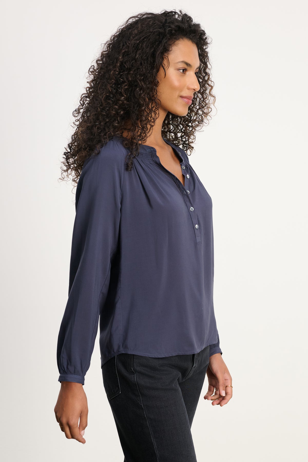 Woman with curly hair wears the AURELIA TOP by Velvet by Graham & Spencer—a navy blue rayon challis top with a smocked neckline—paired with black pants, standing in profile against a plain light background.-39800290508993