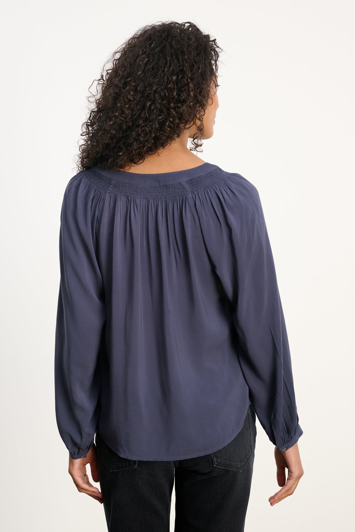 A person with curly hair is shown from the back against a plain background, wearing the Velvet by Graham & Spencer AURELIA TOP—a long-sleeve blouse with a smocked neckline—paired with black pants.-39800290476225