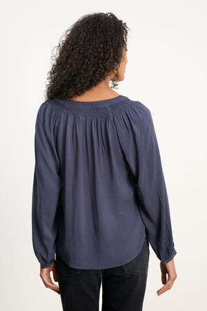 A person with curly hair is shown from the back against a plain background, wearing the Velvet by Graham & Spencer AURELIA TOP—a long-sleeve blouse with a smocked neckline—paired with black pants.