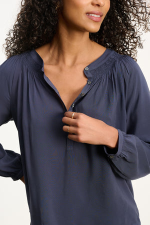 A woman with curly hair wears the AURELIA TOP by Velvet by Graham & Spencer in navy blue rayon challis, featuring a partially unbuttoned collar, and holds her hand to her chest, displaying a simple gold ring.