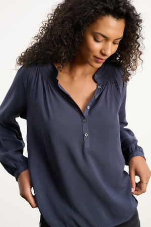 A woman with curly hair wears the AURELIA TOP by Velvet by Graham & Spencer, a navy blue long sleeve blouse with a smocked neckline, looking down with her hands near her hips.