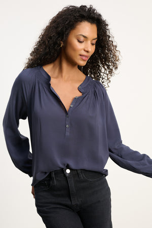 A woman with curly hair wears the AURELIA TOP by Velvet by Graham & Spencer—navy rayon challis, long sleeves—paired with black jeans; she looks down with a slight smile against a plain background.