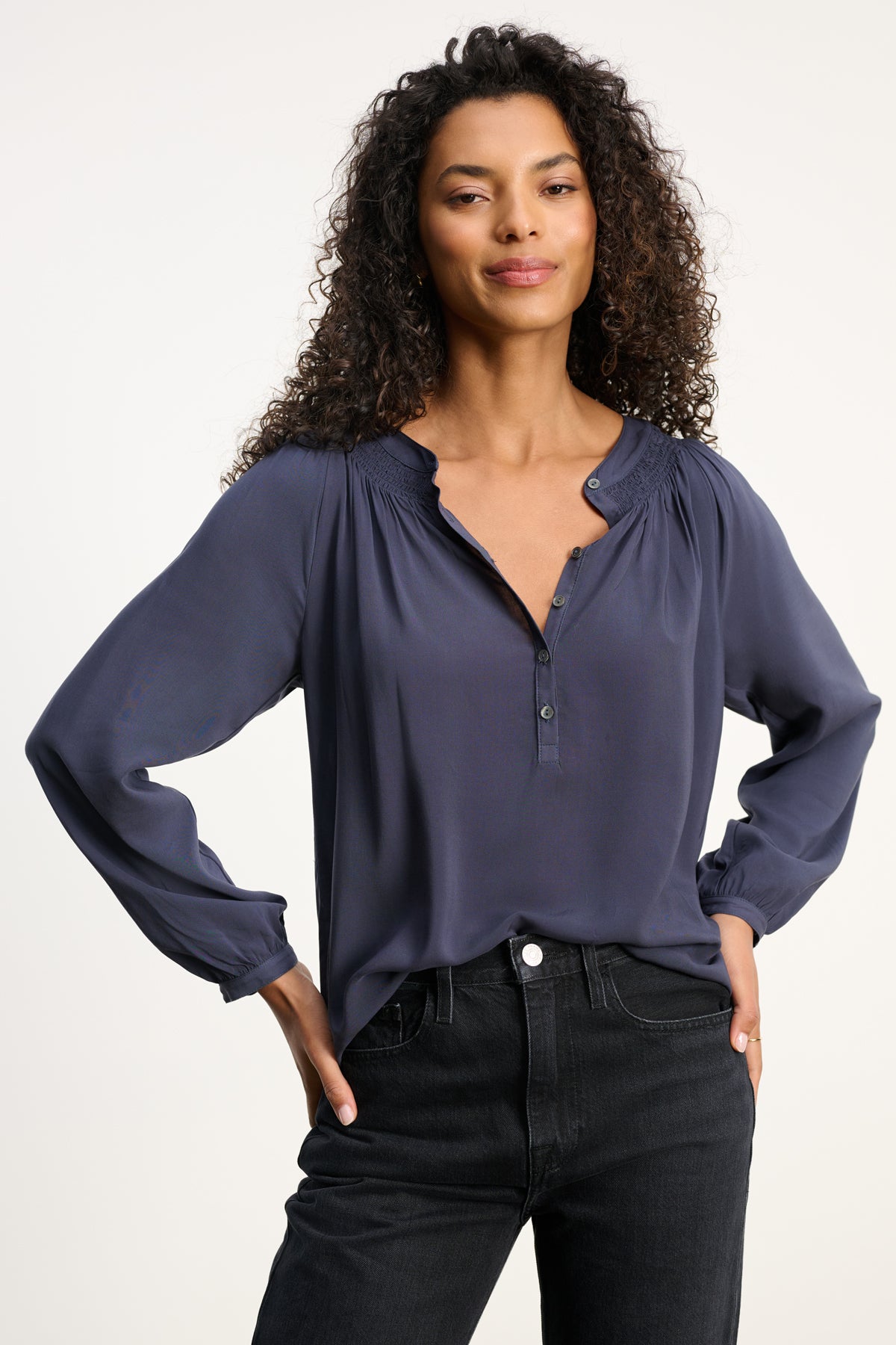 A woman stands facing forward against a plain light background, wearing the Velvet by Graham & Spencer AURELIA TOP—a long sleeve dark blue blouse with a smocked neckline—paired with black jeans.-39800290345153