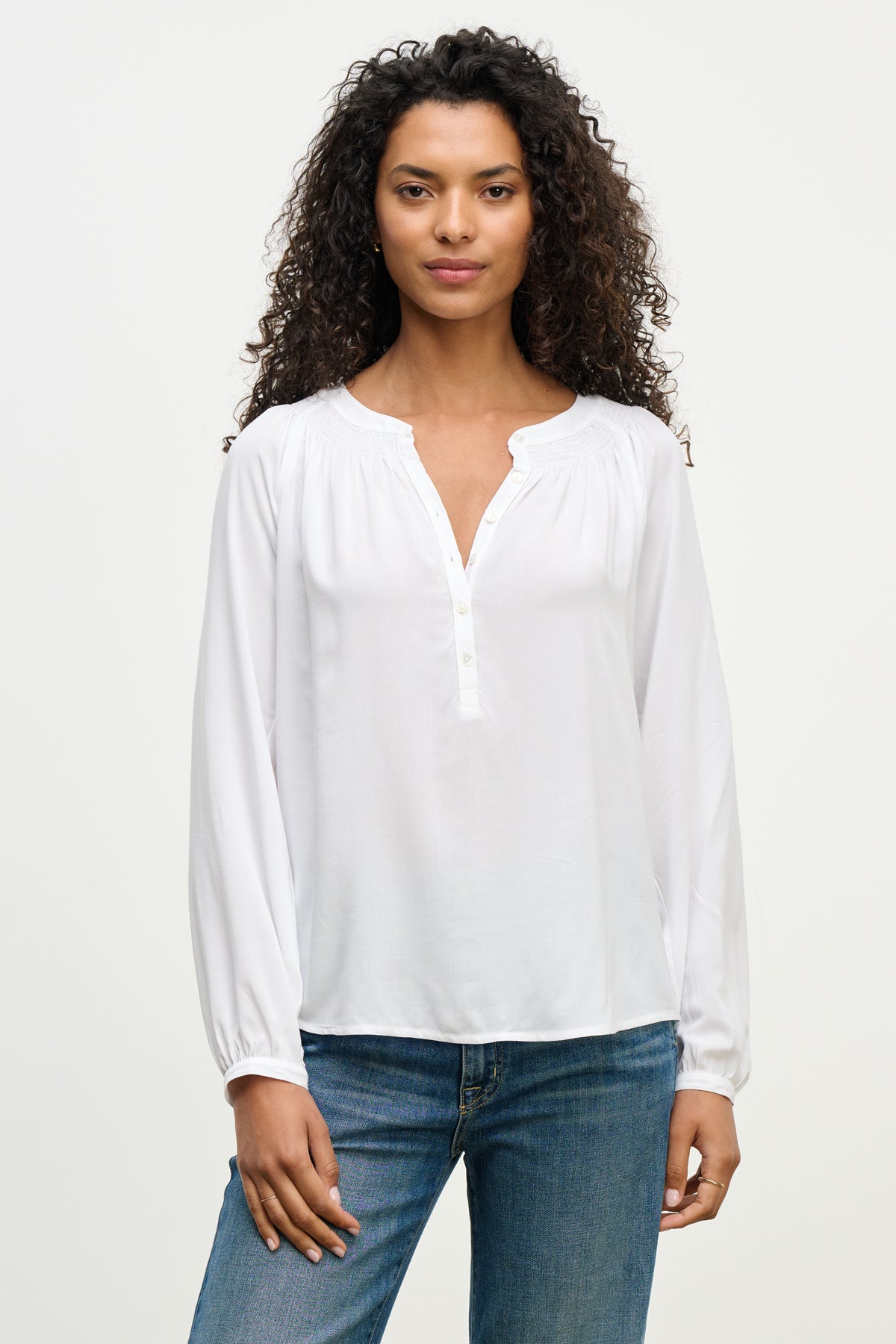 A woman stands against a plain background wearing the AURELIA TOP by Velvet by Graham & Spencer, a long sleeve breathable rayon challis top with a buttoned neckline, paired with blue jeans.-40227028730049