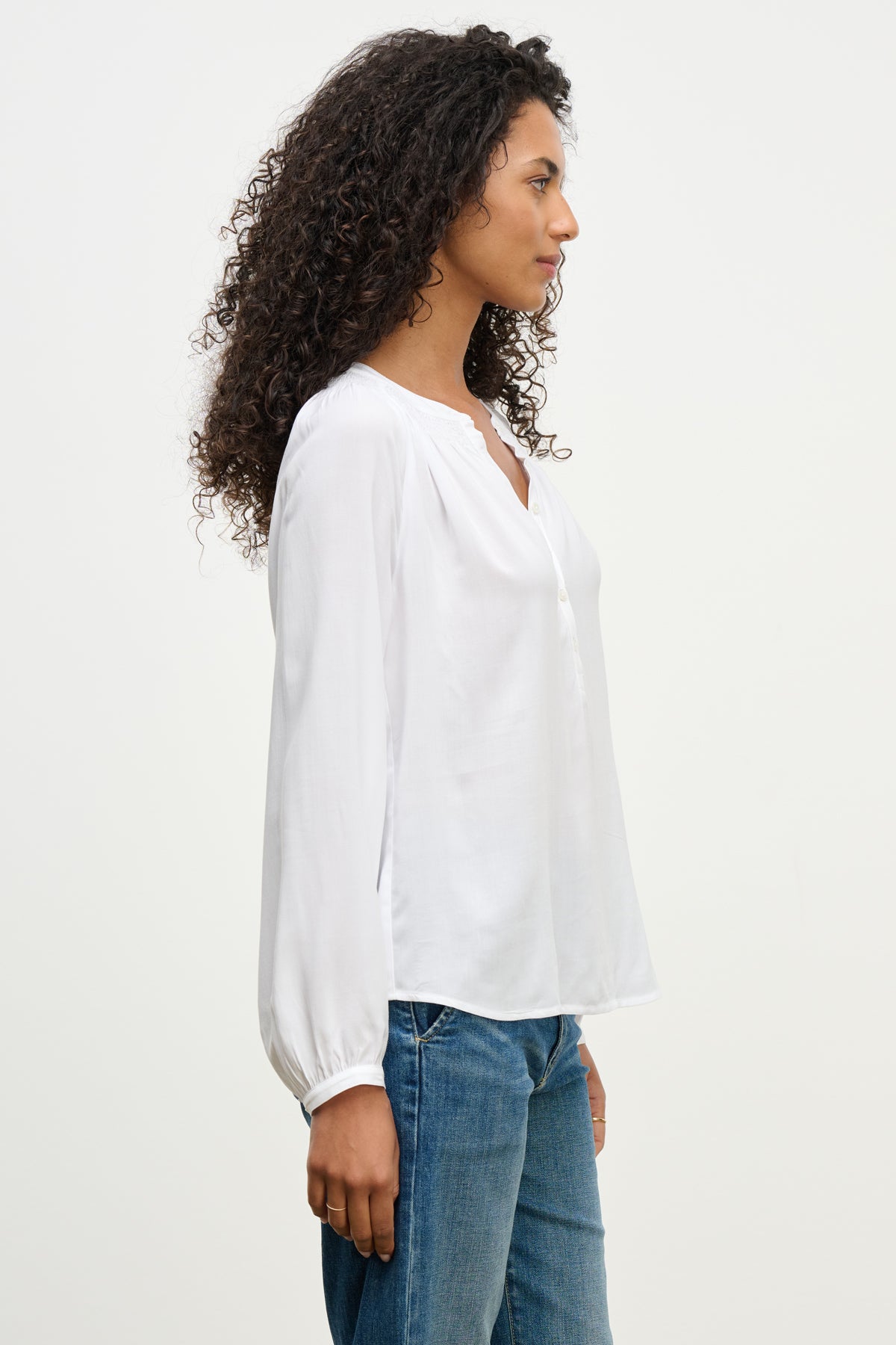 A woman with curly hair stands in profile against a plain light background, wearing the AURELIA TOP by Velvet by Graham & Spencer—a white, long-sleeve top with smocked detailing—paired with blue jeans.