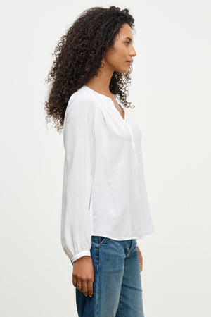 A woman with curly hair stands in profile against a plain light background, wearing the AURELIA TOP by Velvet by Graham & Spencer—a white, long-sleeve top with smocked detailing—paired with blue jeans.