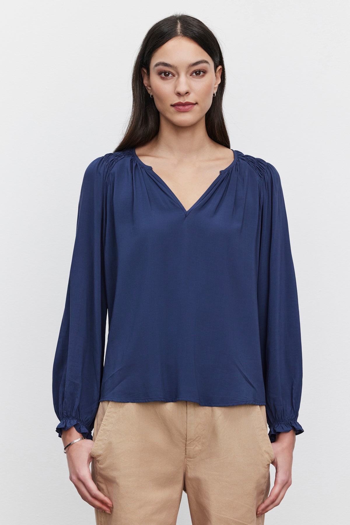   A woman with long dark hair wears a blue AZALEA BLOUSE by Velvet by Graham & Spencer, featuring a v-neckline and elastic ruffle cuffs, paired with beige pants, standing against a plain background. 