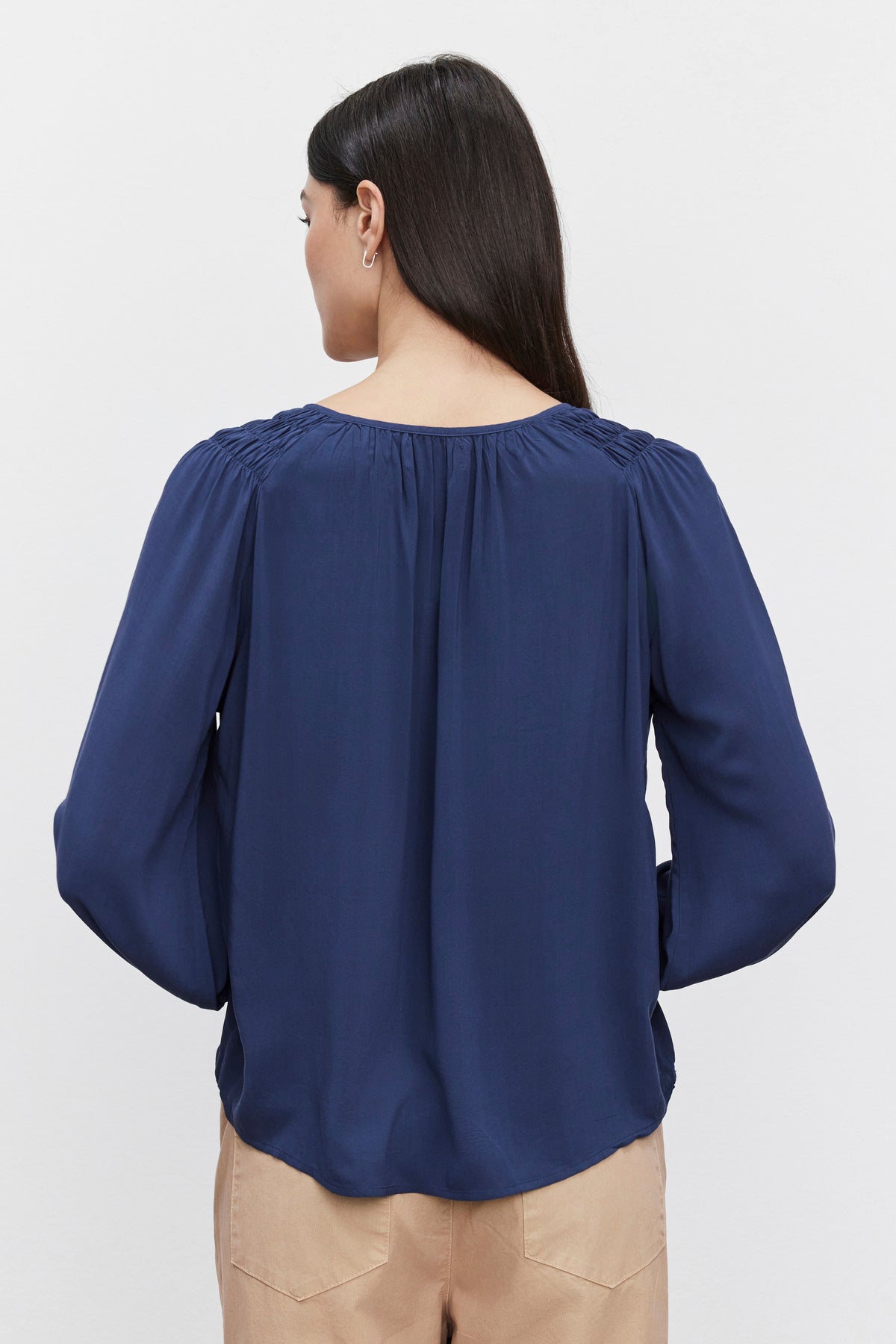   Person with long dark hair wearing a Velvet by Graham & Spencer AZALEA BLOUSE with elastic ruffle cuffs and beige pants, photographed from the back against a plain white background. 