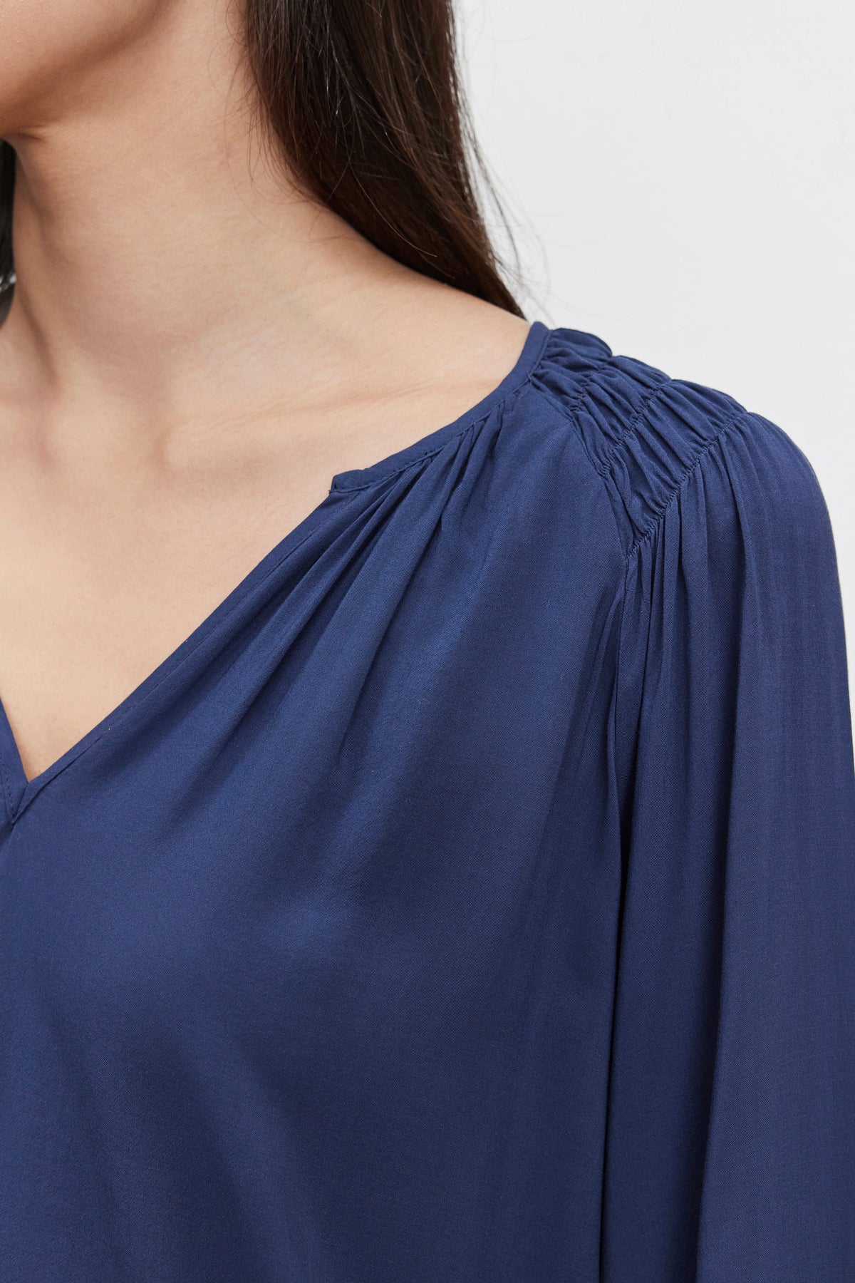   Close-up of a person's upper body wearing the AZALEA BLOUSE by Velvet by Graham & Spencer in dark blue with a V-neckline. Their head is slightly turned to the right, and their long hair cascades over their shoulder. The elastic ruffle cuffs add a touch of elegance to the ensemble. 