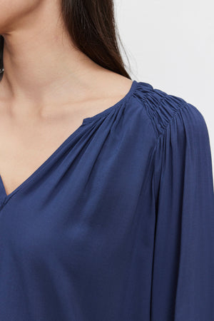 Close-up of a person's upper body wearing the AZALEA BLOUSE by Velvet by Graham & Spencer in dark blue with a V-neckline. Their head is slightly turned to the right, and their long hair cascades over their shoulder. The elastic ruffle cuffs add a touch of elegance to the ensemble.