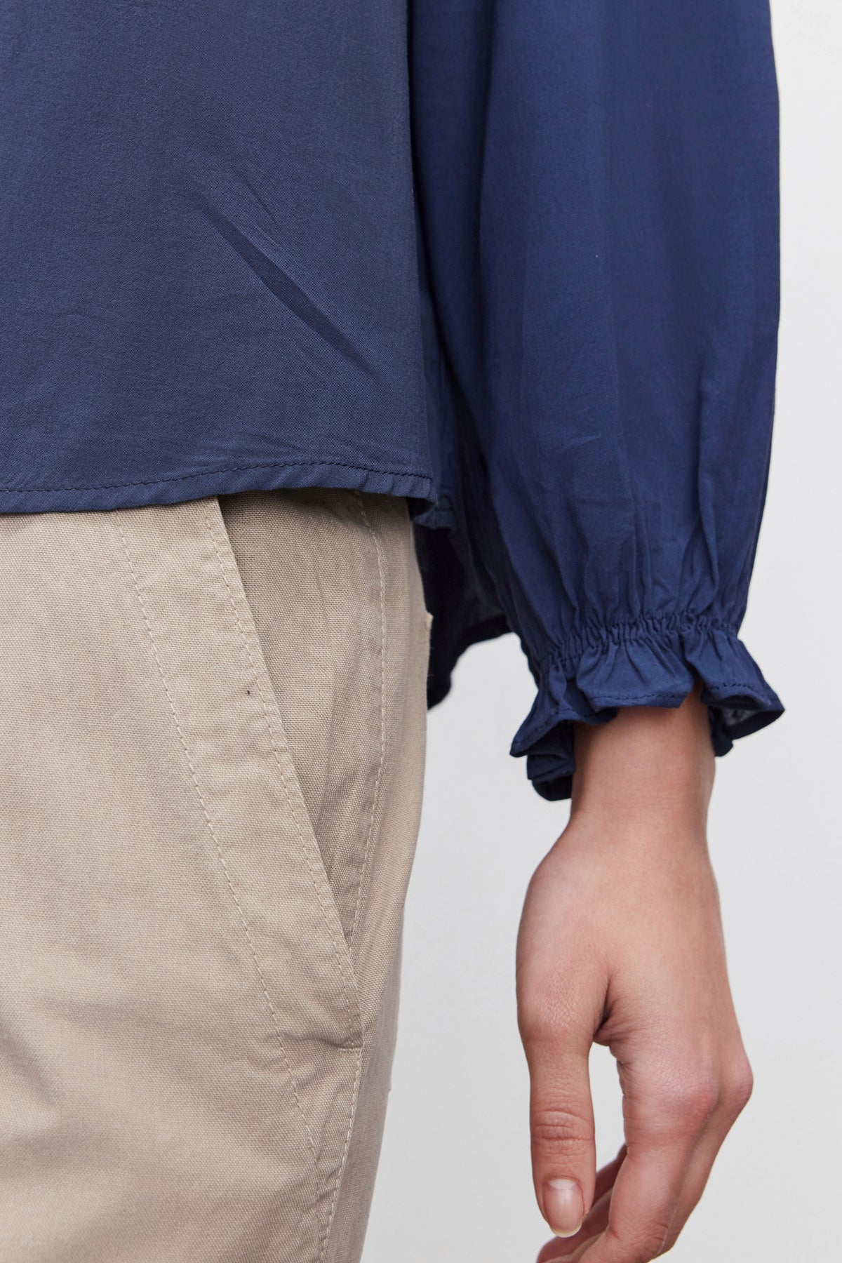   A close-up of a person wearing the AZALEA BLOUSE by Velvet by Graham & Spencer, showcasing its V-neckline and elastic ruffle cuffs, paired with beige pants. The person's hand is relaxed by their side. 