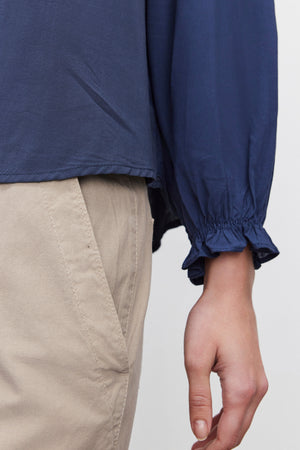 A close-up of a person wearing the AZALEA BLOUSE by Velvet by Graham & Spencer, showcasing its V-neckline and elastic ruffle cuffs, paired with beige pants. The person's hand is relaxed by their side.