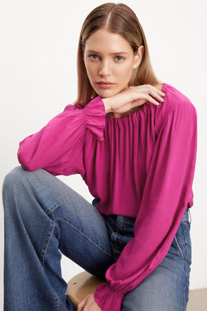 The model is wearing a luxurious Velvet by Graham & Spencer pink Bristol Neck Tie Top made of rayon challis fabric and paired with jeans.