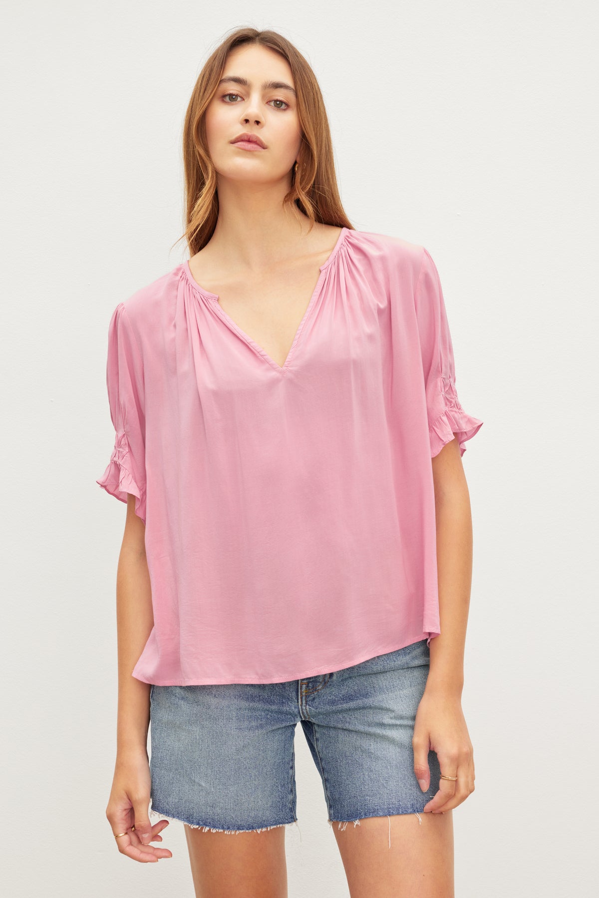 Woman in a CALISSA SPLIT NECK BLOUSE by Velvet by Graham & Spencer with shirred detailing and denim shorts standing against a neutral background.-36387174482113