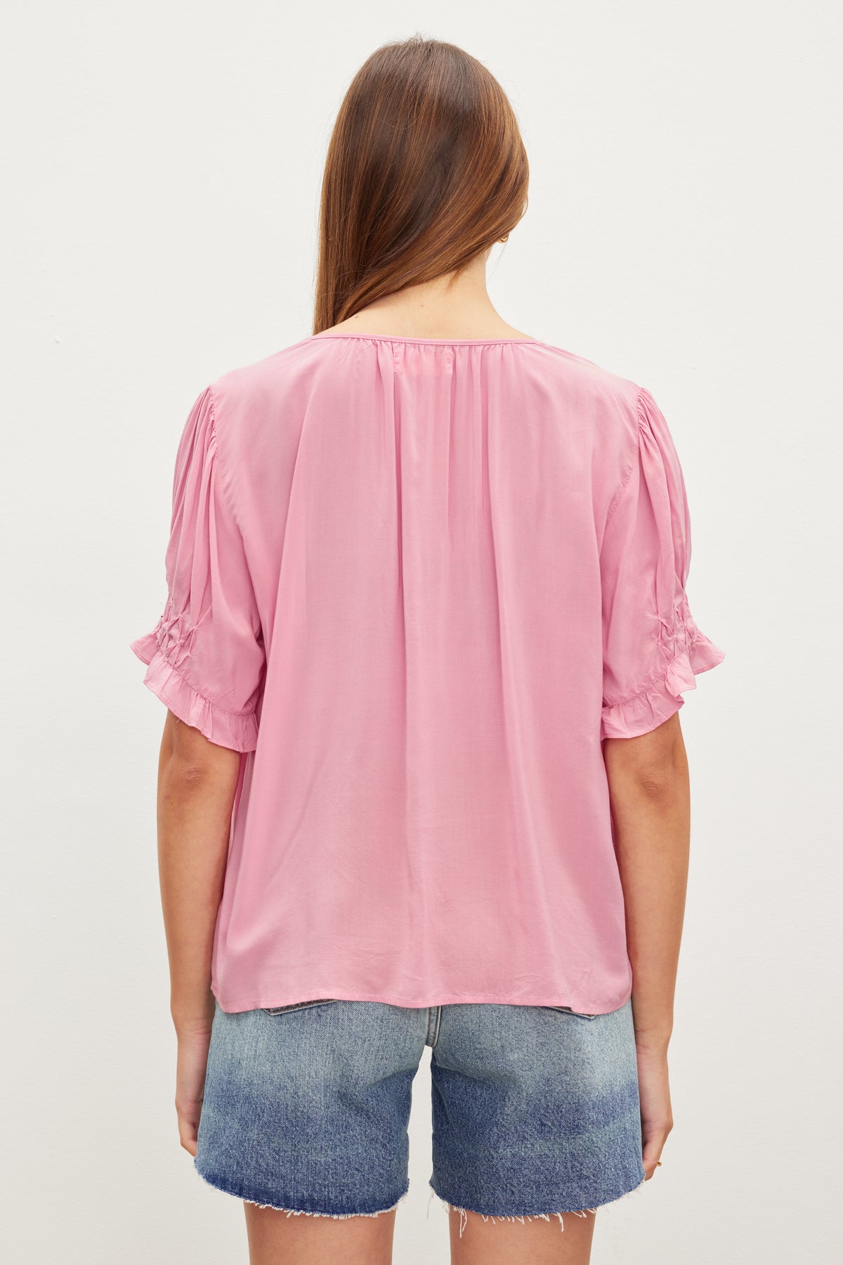Woman standing with her back to the camera, wearing a pink Velvet by Graham & Spencer Calissa Split Neck Blouse and denim shorts.-36387174547649