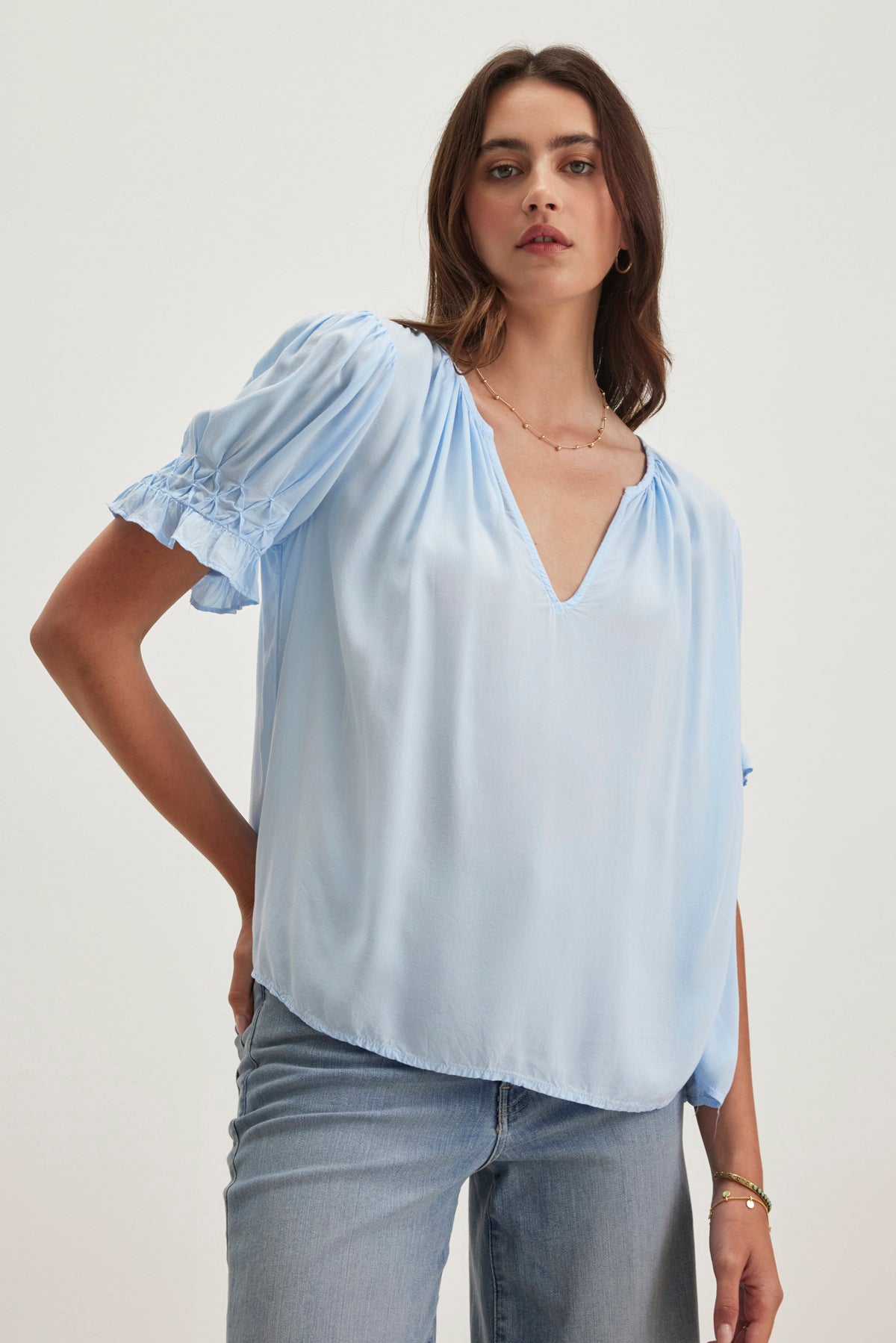A woman models the CALISSA SPLIT NECK BLOUSE from Velvet by Graham & Spencer, a relaxed-fit light blue rayon challis top, pairing it perfectly with jeans against a plain background.-38918675235009