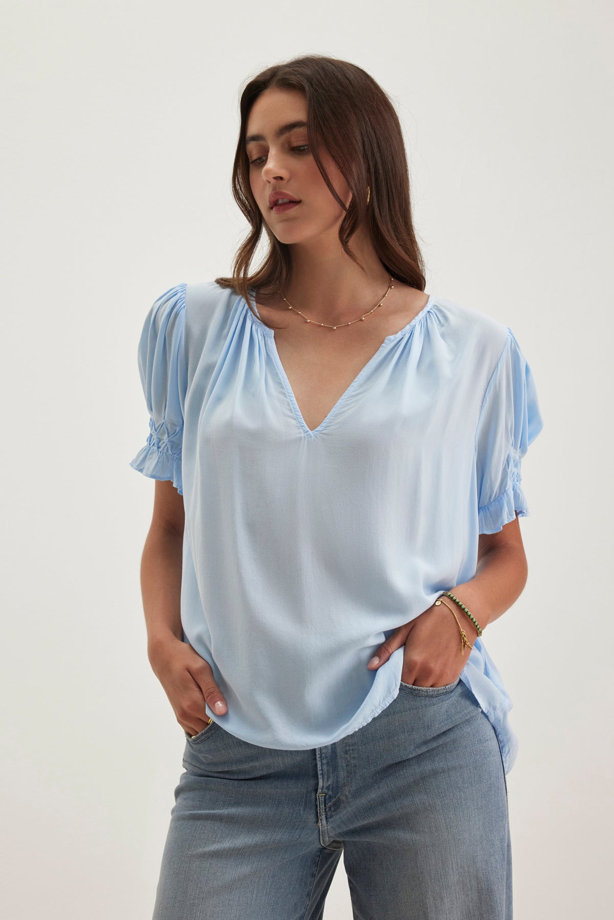 A person with long hair stands wearing a Velvet by Graham & Spencer CALISSA SPLIT NECK BLOUSE made from soft rayon challis, paired with jeans. Their hands rest in their pockets against a plain background.-38918675497153