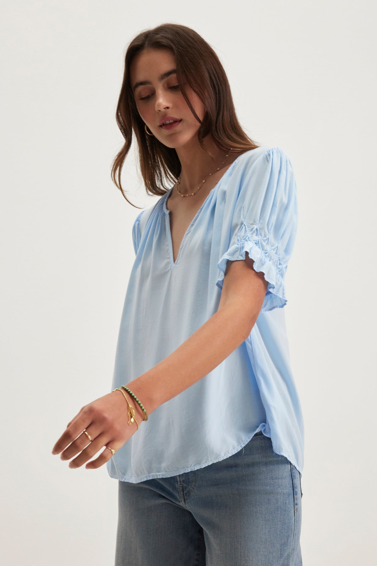 Someone is wearing the CALISSA SPLIT NECK BLOUSE by Velvet by Graham & Spencer, a relaxed fit light blue top made from rayon challis with short ruched sleeves, paired stylishly with jeans against a plain background.-38918675136705