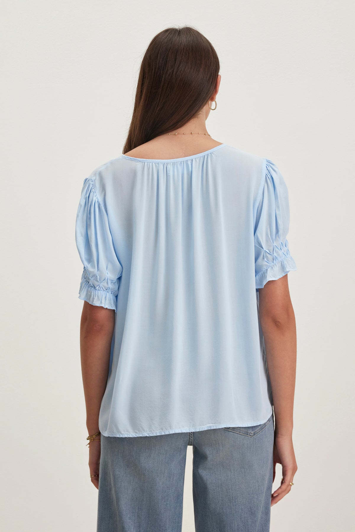 A woman with long dark hair faces away, wearing the CALISSA SPLIT NECK BLOUSE by Velvet by Graham & Spencer, made of soft rayon challis, paired perfectly with her blue jeans.-38918675071169