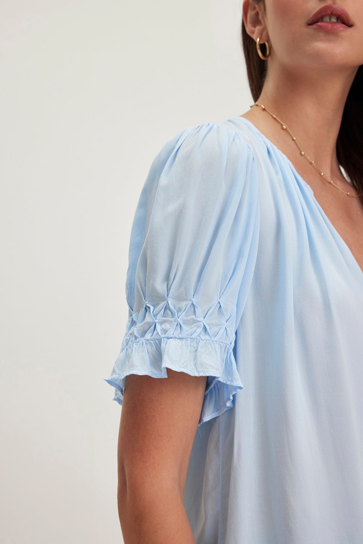 A person styled in Velvet by Graham & Spencer's CALISSA SPLIT NECK BLOUSE—a light blue rayon challis top with puffed sleeves and ruffle details—paired with a gold necklace and hoop earrings.-38918675202241