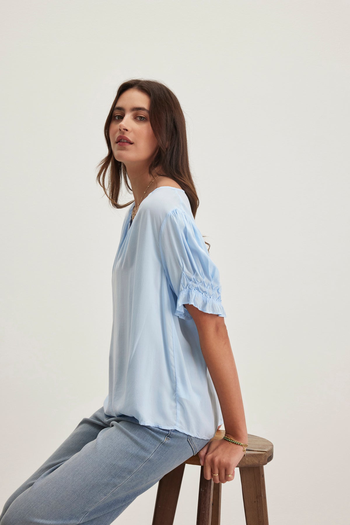 A woman wearing a Velvet by Graham & Spencer CALISSA SPLIT NECK BLOUSE, featuring a relaxed fit and light blue color, paired with jeans, sits on a wooden stool against a plain background.-38918675464385