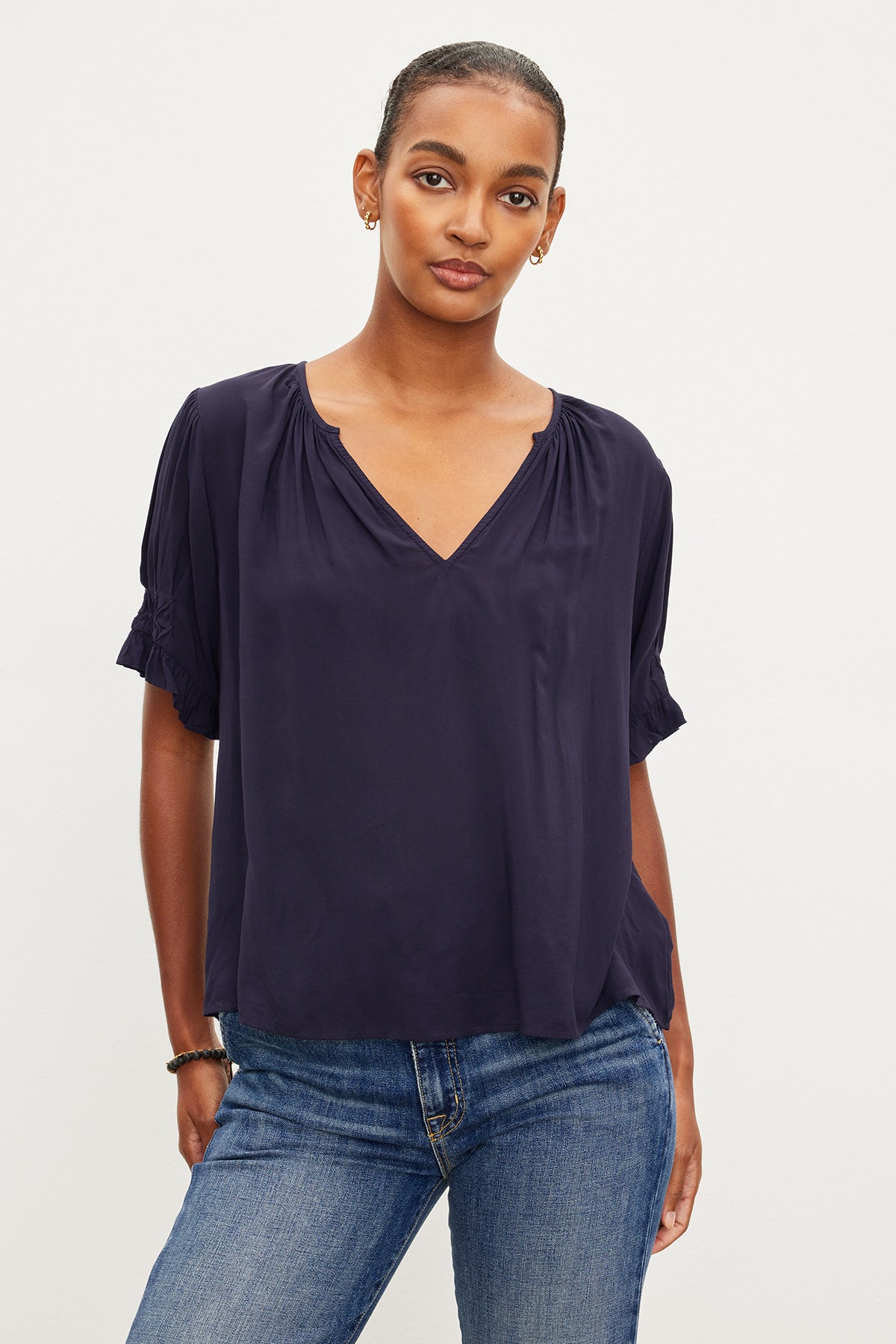 A person standing, wearing a navy blue Velvet by Graham & Spencer CALISSA SPLIT NECK BLOUSE with ruffle cuffs and blue jeans, with a neutral expression on a plain white background.-36909466845377