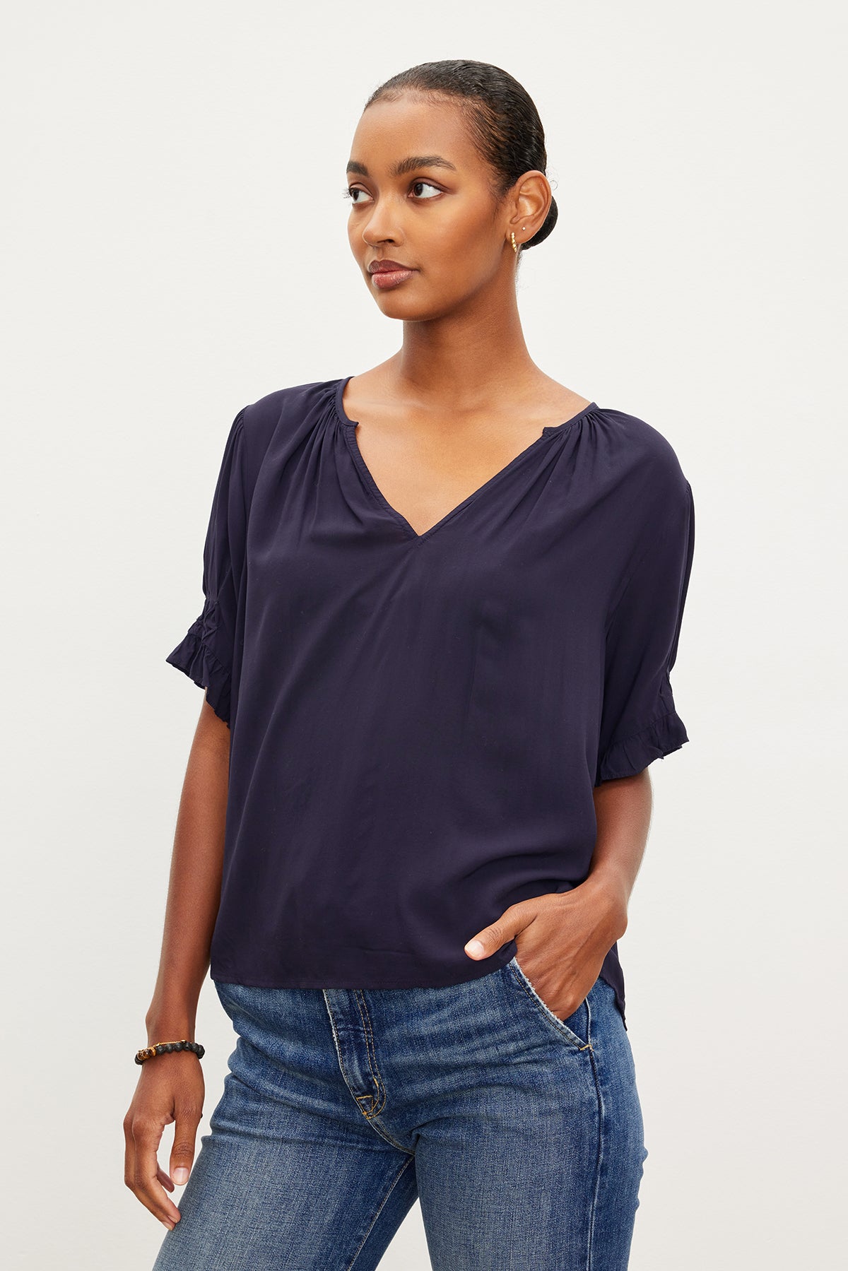   A person with short hair wears a dark blue Velvet by Graham & Spencer CALISSA SPLIT NECK BLOUSE with ruffle cuffs and jeans, posing with one hand in their pocket against a plain background. 