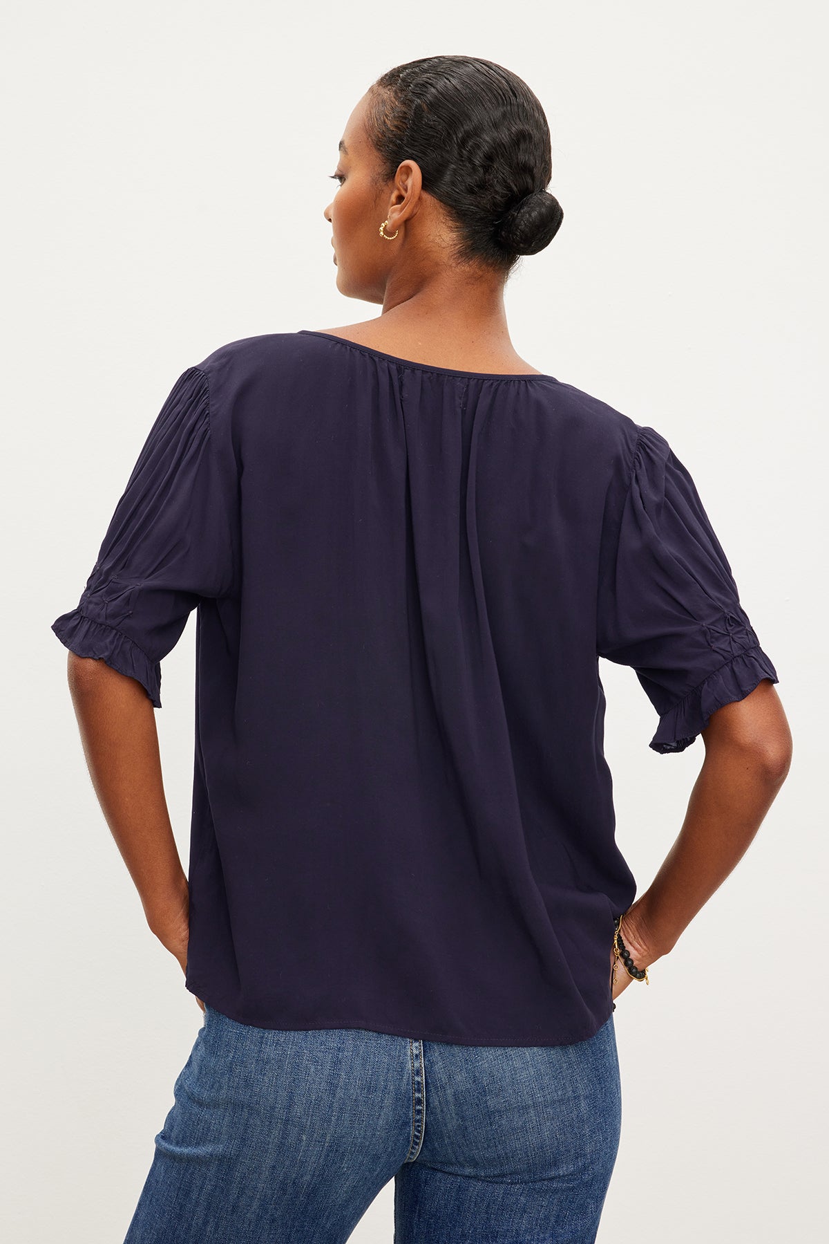   A person stands facing away, wearing a dark blue CALISSA SPLIT NECK BLOUSE by Velvet by Graham & Spencer with short, ruffled sleeves and blue jeans. Their hair is pulled back into a neat bun. 