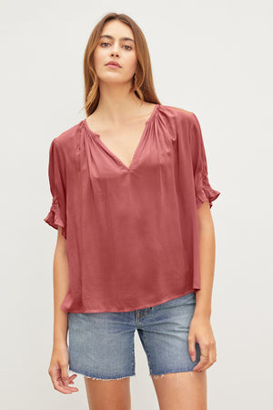 A person with long hair wears a loose-fitting, short-sleeved, deep pink CALISSA SPLIT NECK BLOUSE by Velvet by Graham & Spencer with a relaxed fit and denim shorts.