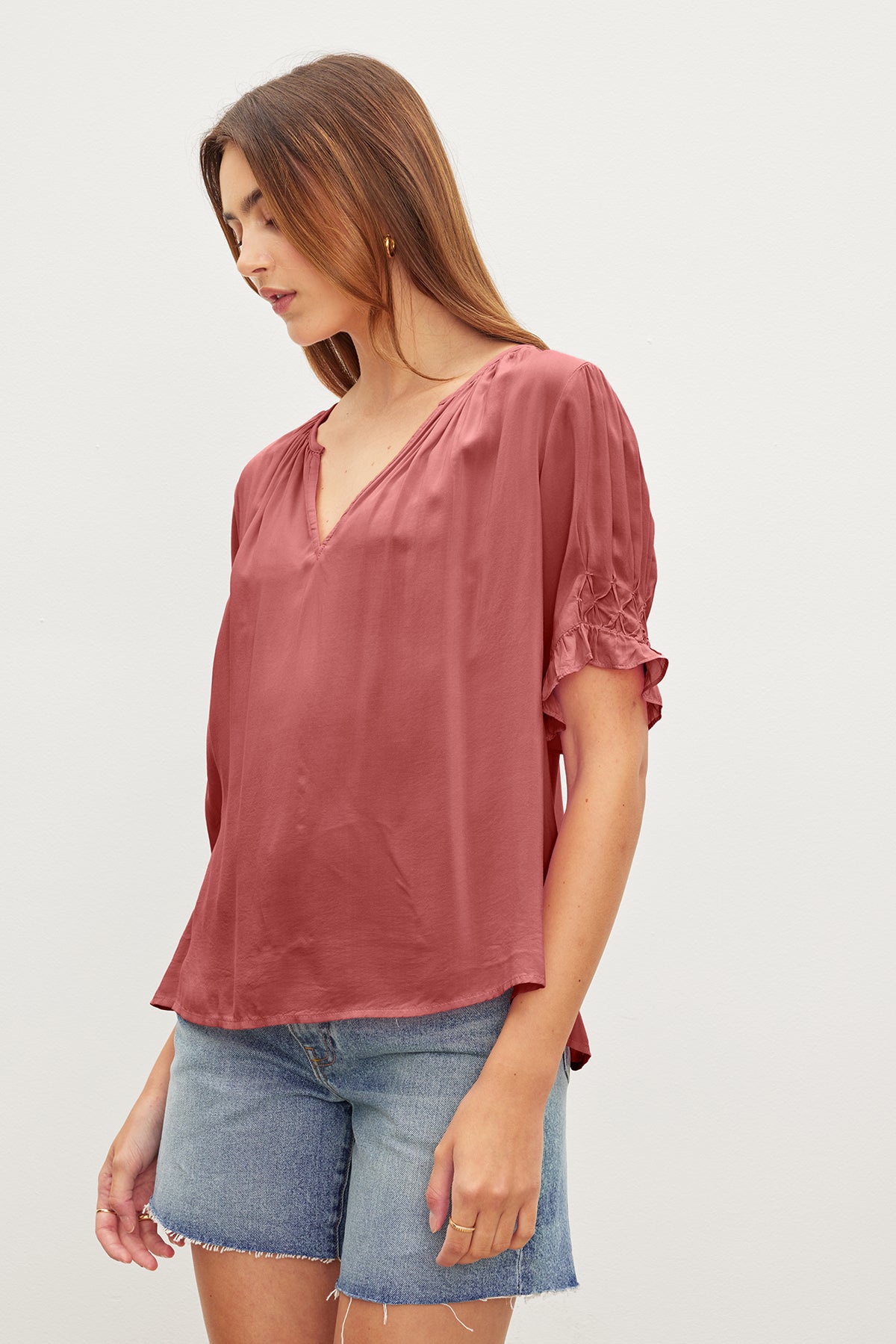 A woman with long brown hair, wearing a relaxed fit pink CALISSA SPLIT NECK BLOUSE by Velvet by Graham & Spencer with ruffle cuffs and denim shorts, stands against a plain white background.-36909459177665