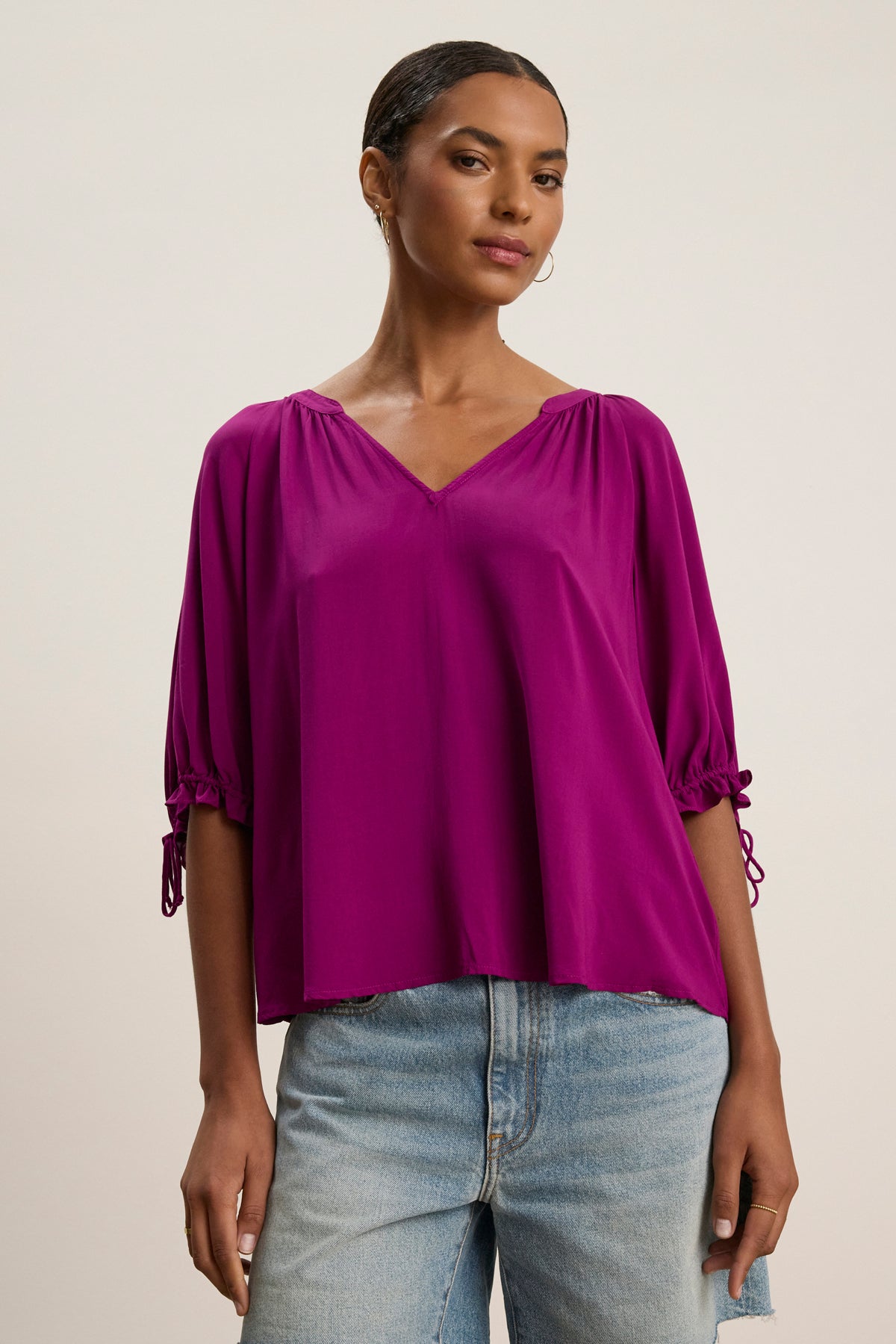 A woman models the Velvet by Graham & Spencer LUNA TOP—a bright purple, relaxed-fit V-neck with short gathered sleeves in soft Rayon Challis—paired with light blue jeans against a plain light background.-39111852818625
