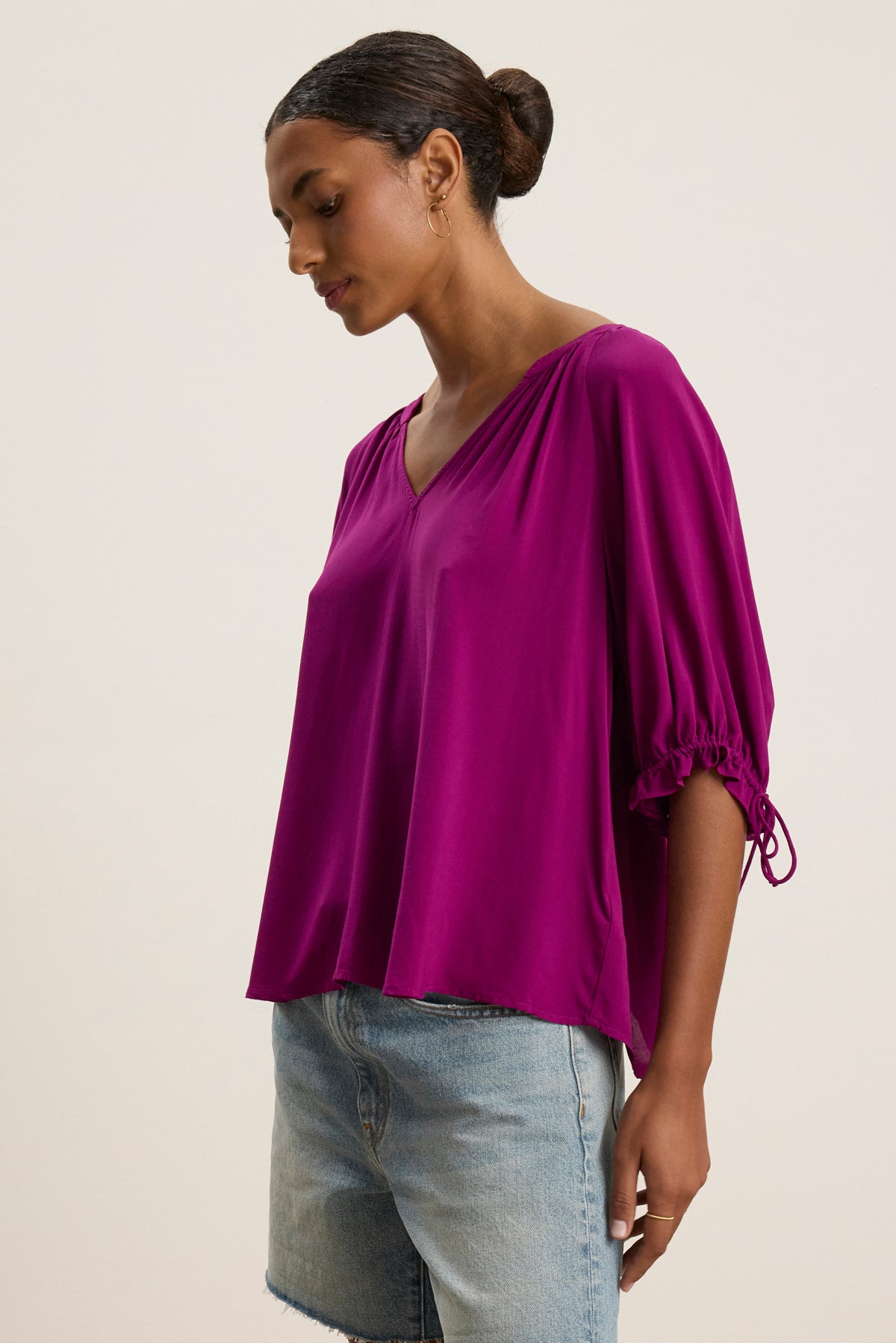 A woman stands against a plain background wearing the LUNA TOP by Velvet by Graham & Spencer, a loose-fitting bright purple V-neck blouse with gathered sleeves, paired with light blue denim shorts.-39111852589249