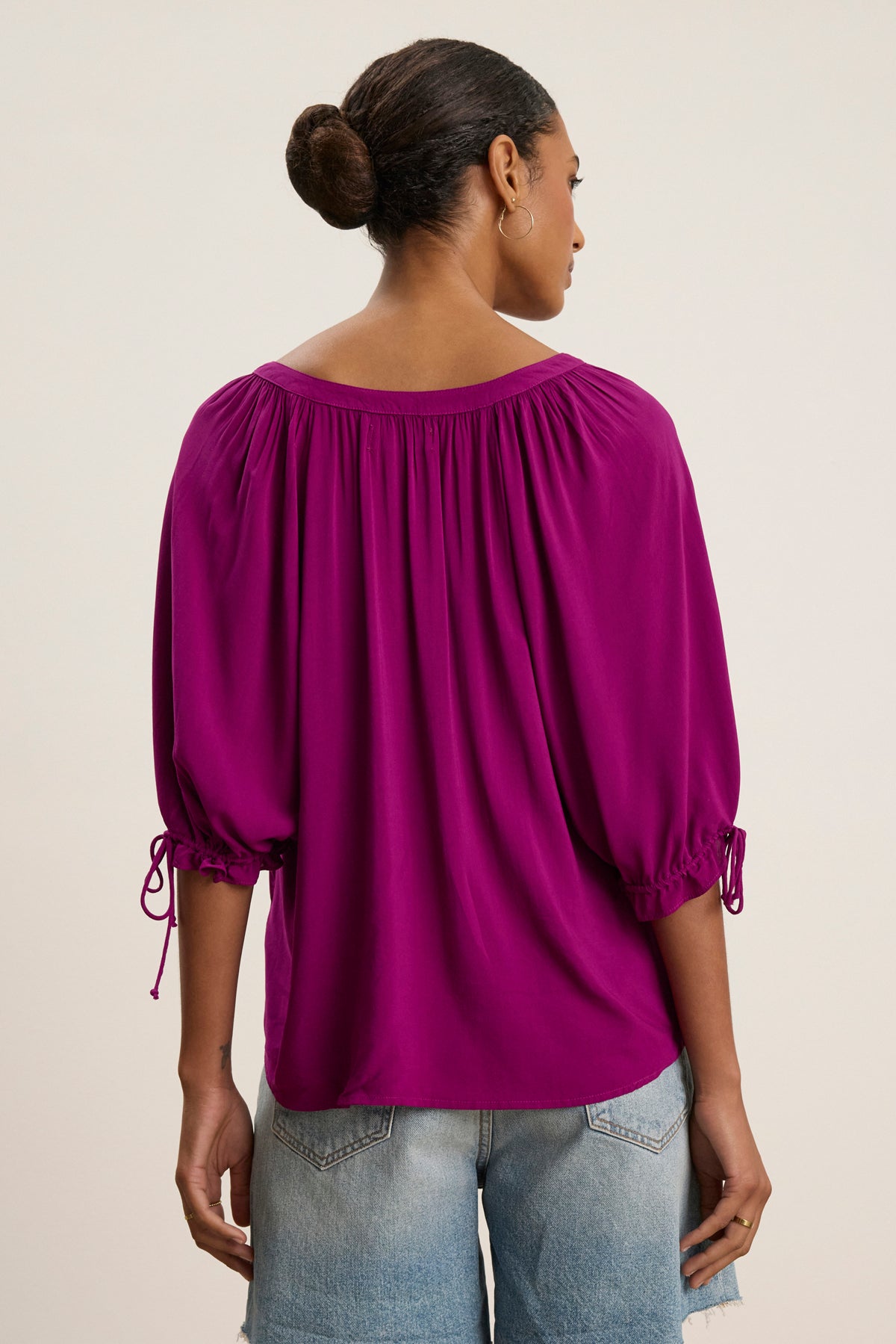 A person faces away, wearing the LUNA TOP by Velvet by Graham & Spencer—a bright magenta rayon challis blouse with gathered sleeves—paired with light blue denim shorts, set against a neutral background.-39111851573441