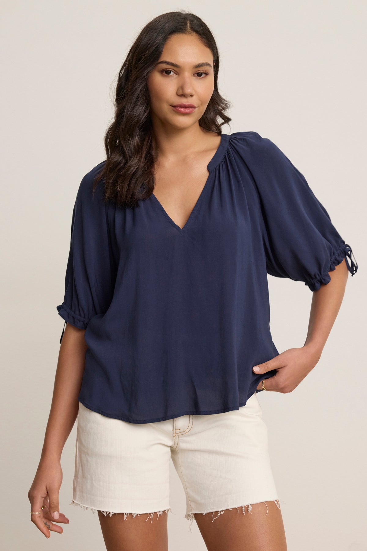 A woman stands against a plain background, wearing the LUNA TOP by Velvet by Graham & Spencer—a navy V-neck with elbow-length sleeves in soft Rayon Challis—paired with white frayed hem shorts.-39148730482881