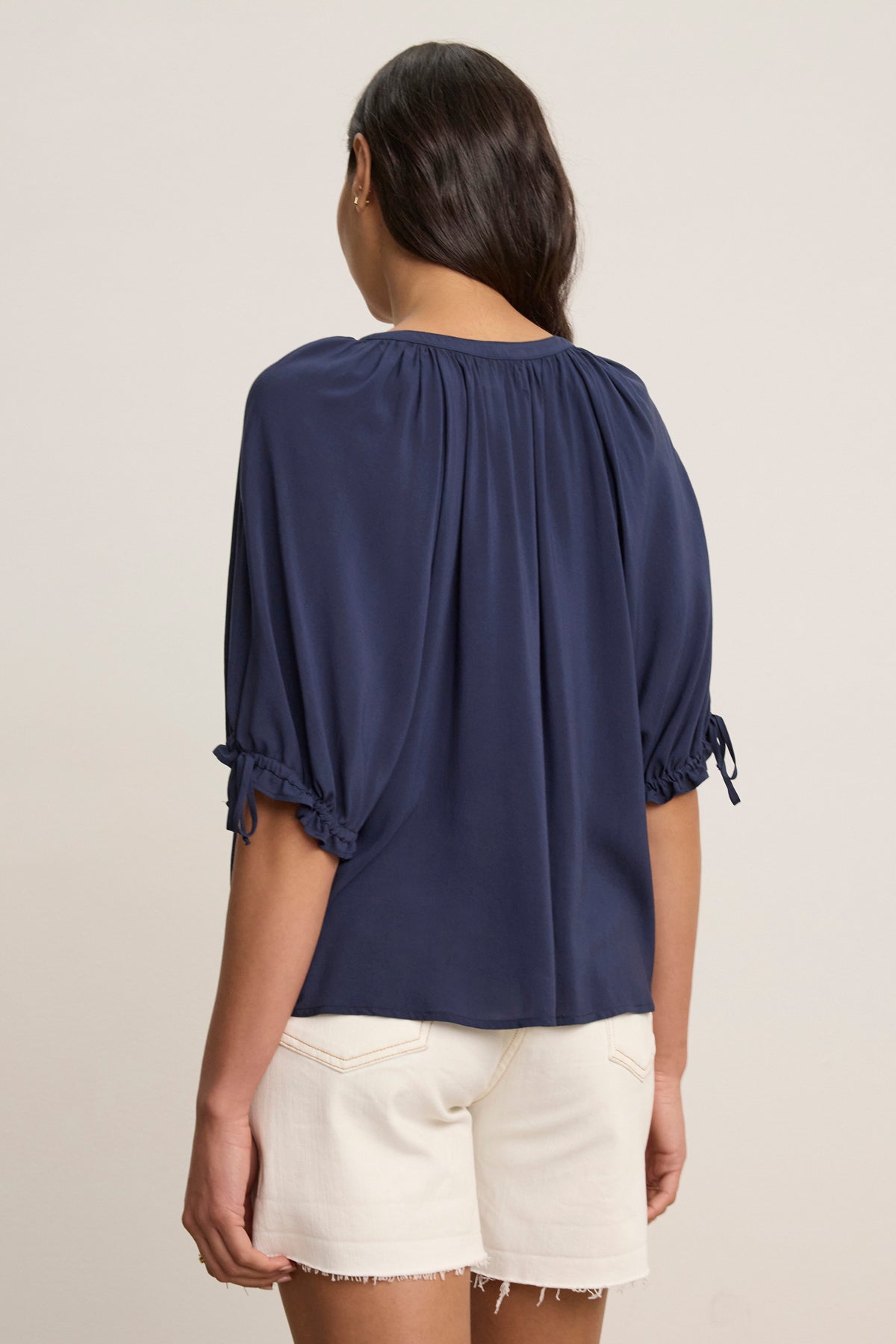 A woman with long dark hair, seen from the back, wears the Velvet by Graham & Spencer LUNA TOP— a versatile navy Rayon Challis V-neck with elbow-length sleeves—paired with white frayed hem shorts.-39148730417345