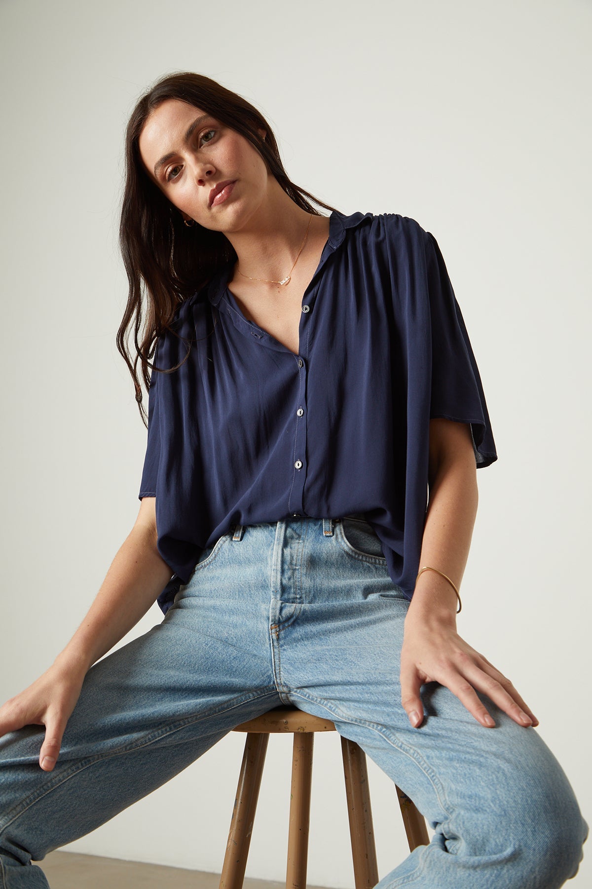 A woman is sitting on a stool wearing jeans and a Velvet by Graham & Spencer Nikki Button-Up Top.-26496273088705
