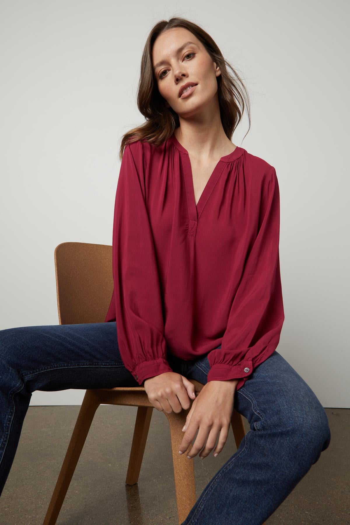 A woman is sitting on a chair wearing jeans and a Velvet by Graham & Spencer POSIE SPLIT NECK BLOUSE.-26861471105217